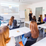 Un'aula luminosa in cui si svolge una lezione di formazione continua: persone sedute ai tavoli ascoltano un'insegnante davanti a uno schermo di proiezione e una lavagna bianca.