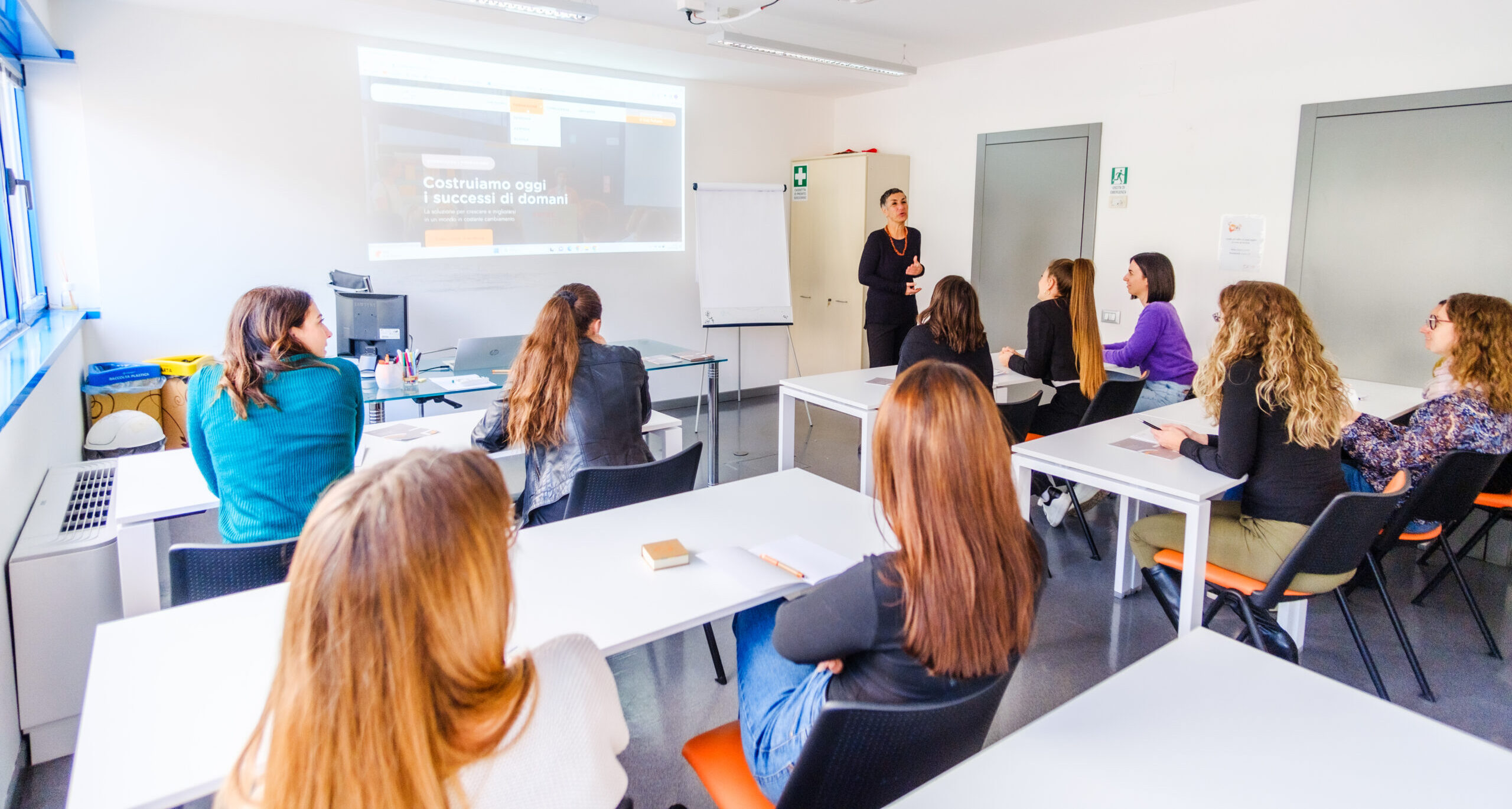 Un'aula luminosa in cui si svolge una lezione di formazione continua: persone sedute ai tavoli ascoltano un'insegnante davanti a uno schermo di proiezione e una lavagna bianca.